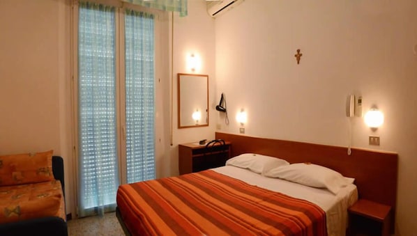 A hotel room with a bed, a nightstand, a desk with a lamp, a chair, and a window with blinds. - Hotel Trinidad (Rimini)