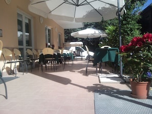 An outdoor patio area with tables and chairs, umbrellas, and a potted plant. - Hotel Trinidad (Rimini)