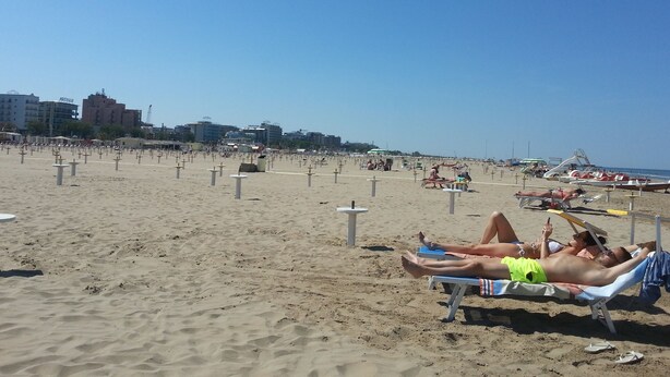 A beach with sun loungers and umbrellas, a clear sky, and buildings in the background.