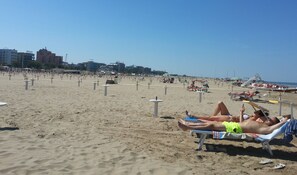 Uma praia com espreguiçadeiras e guarda-sóis, céu claro e edifícios ao fundo.
