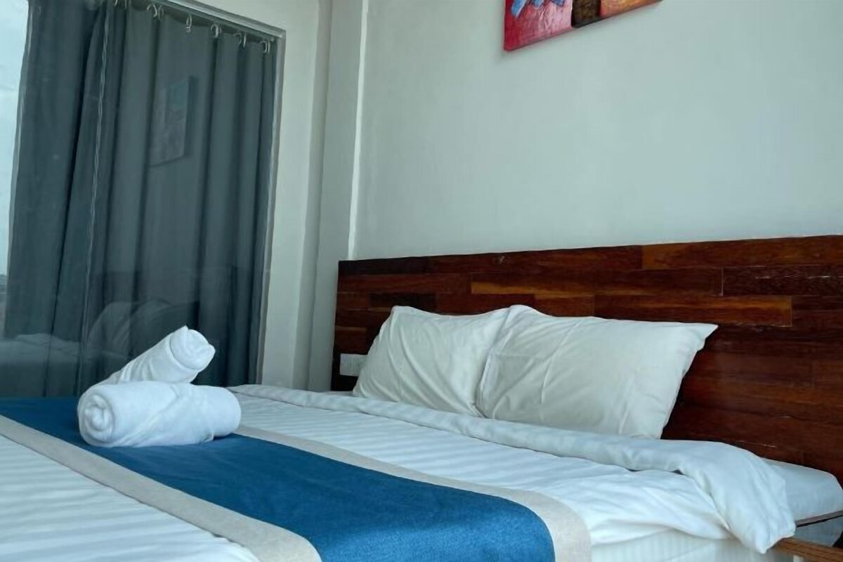 A neatly made bed with white linens and a blue accent, a wooden headboard, and a painting on the wall.
