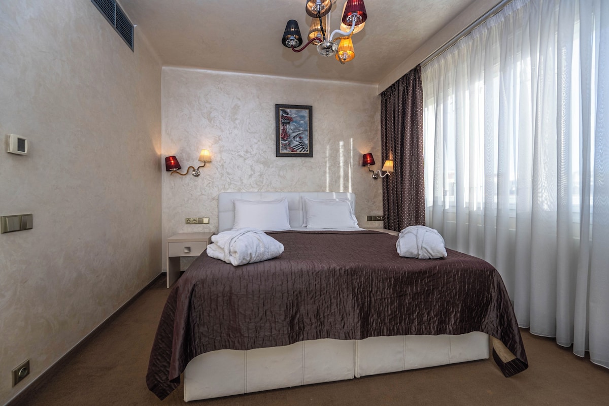A hotel room with a large bed, two bedside tables, a painting on the wall, and a window with curtains.