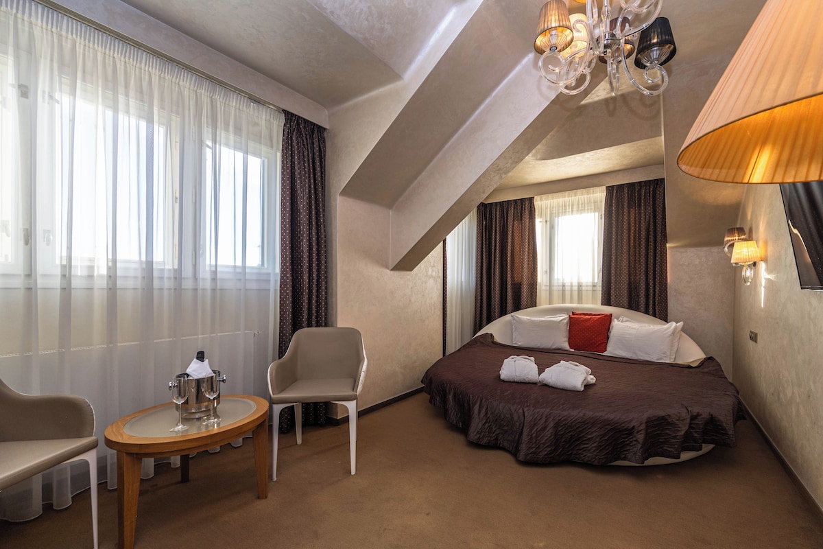 A hotel room with a bed, a chair, a small table with a bottle and glass, and a chandelier.