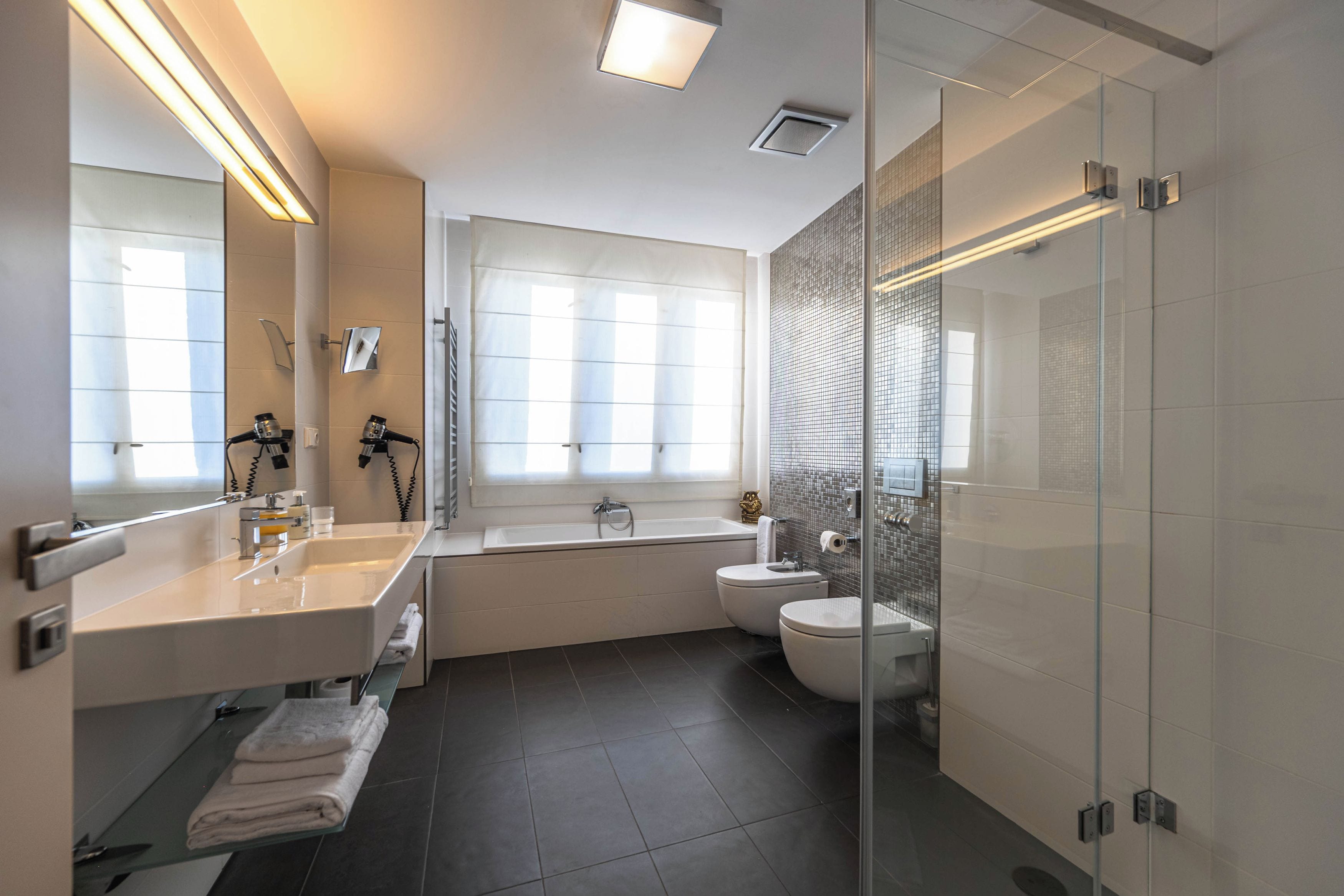A modern bathroom with a large mirror, two sinks, a bathtub, a toilet, and a glass shower enclosure.