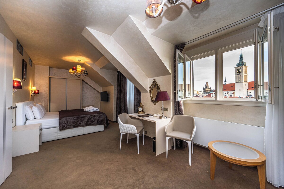 A hotel room with a bed, desk, two chairs, a small table, a window with a city view, and a chandelier.