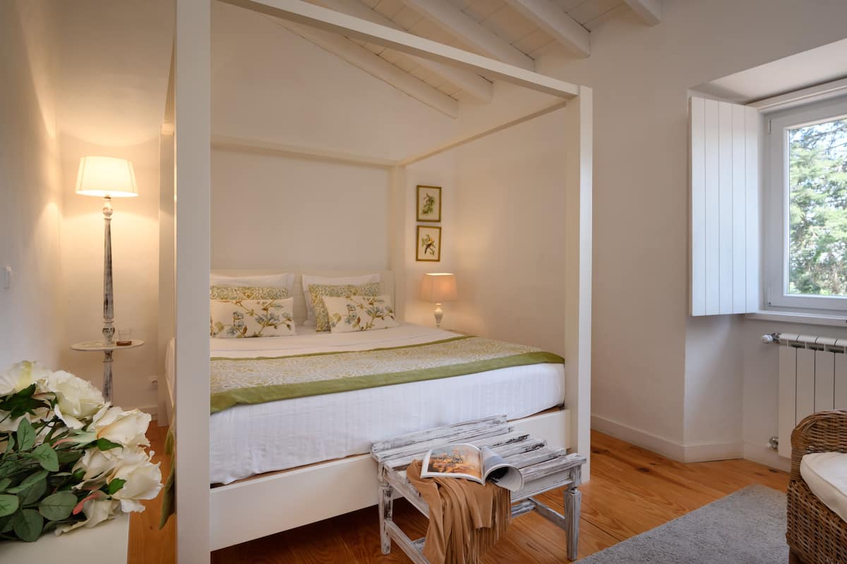 A bedroom with a four-poster bed, a bedside table with a lamp, and a window with a view of trees.