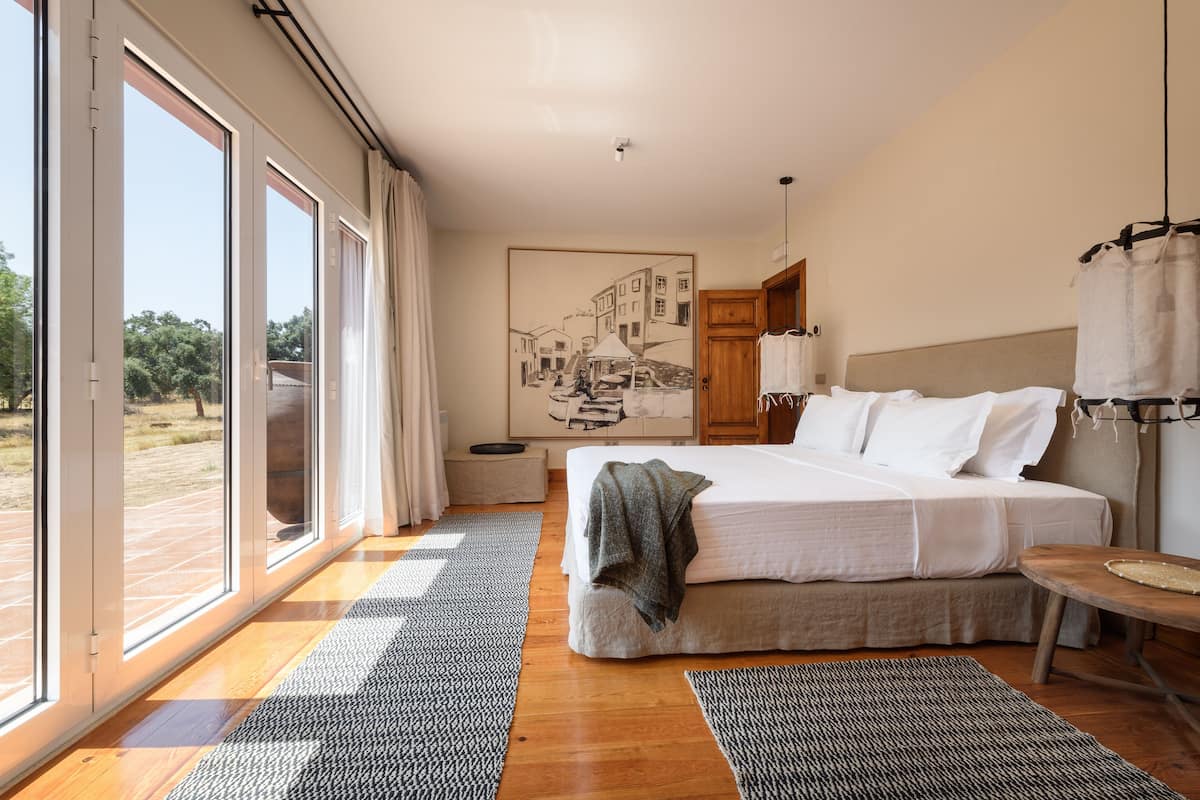 A bedroom with a large bed, a view through the sliding glass doors, a painting on the wall, and a wooden nightstand.