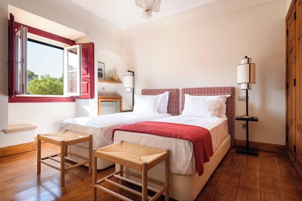 A bedroom with two beds, a window with red shutters, a bedside table, a lamp, and a wooden floor.