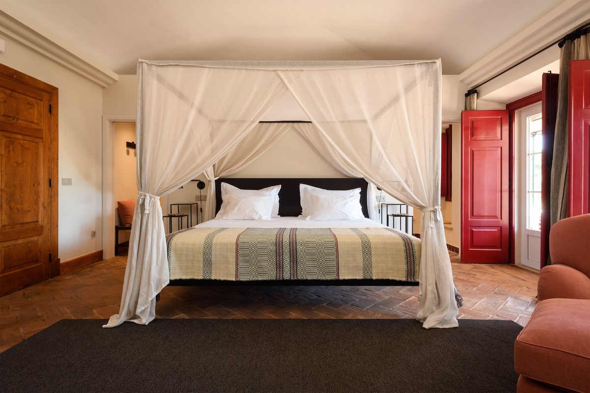 A four-poster canopy bed with a patterned bedspread, a wooden floor, and a red door in the background.
