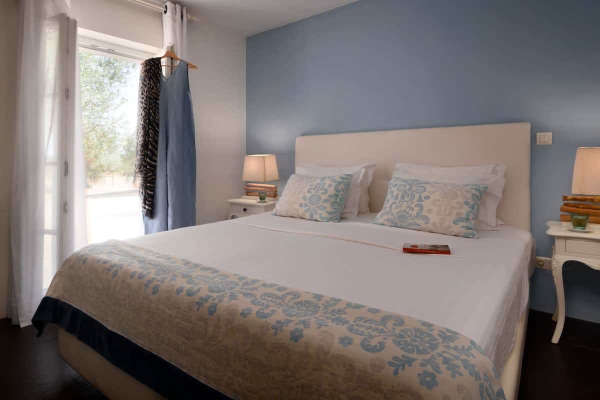 A bedroom with a large bed, two bedside tables with lamps, a window with curtains, and a view of trees outside.