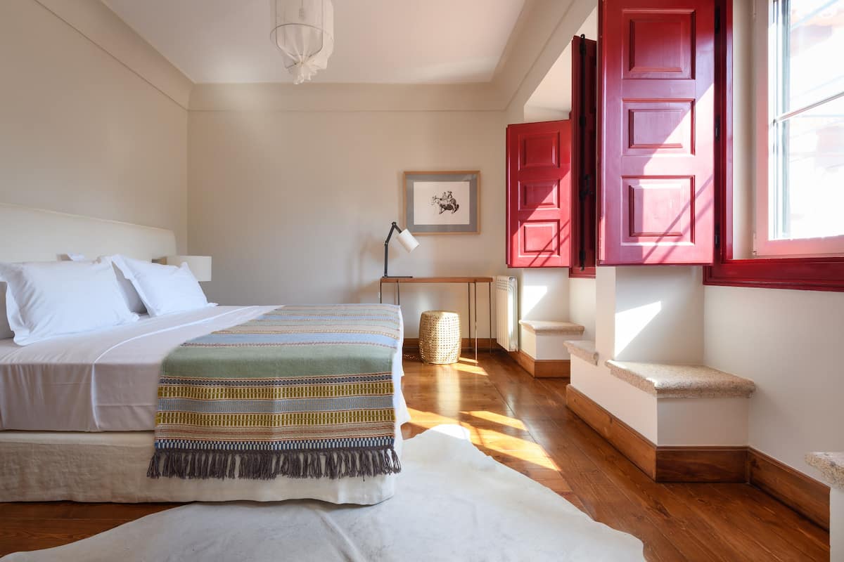 A bedroom with a bed, a bedside table, a lamp, a framed picture, and a window with red shutters.