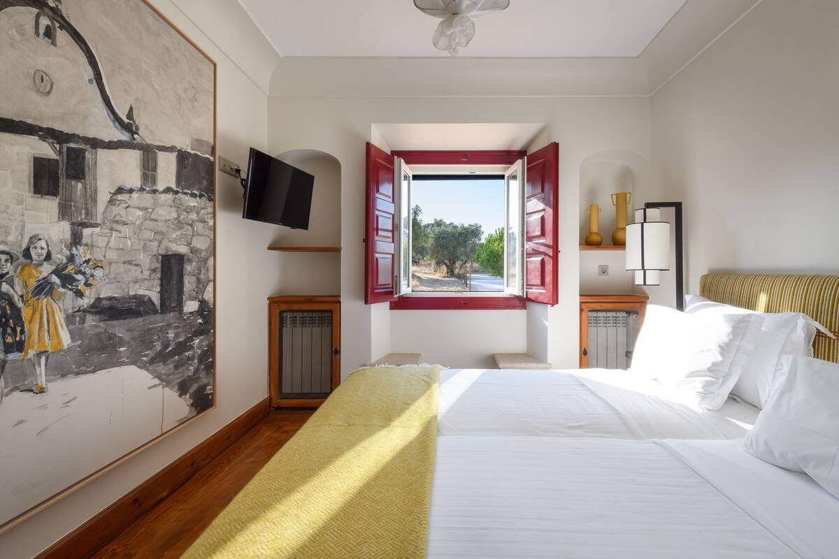 A bedroom with a bed, a painting on the wall, a television, a window with red shutters, and a view of the outside.