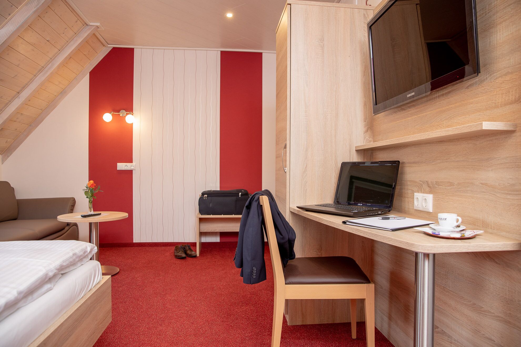 A compact hotel room with a bed, a desk with a laptop, a chair, a small table with a remote control, and a red carpet.