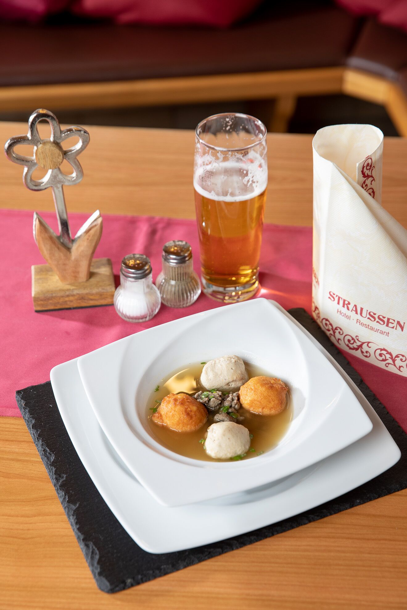 A table setting with a white plate containing a soup dish with dumplings, a glass of beer, and a napkin with a logo.