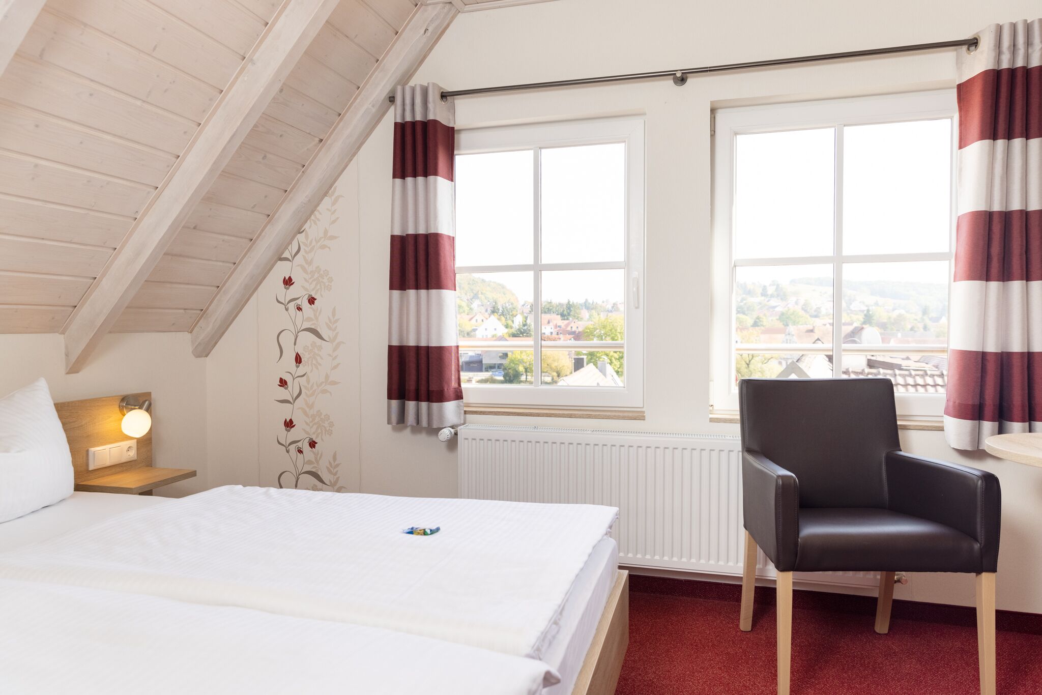 Habitación de hotel con cama, una silla, una mesita de noche con lámpara y ventana con cortinas.
