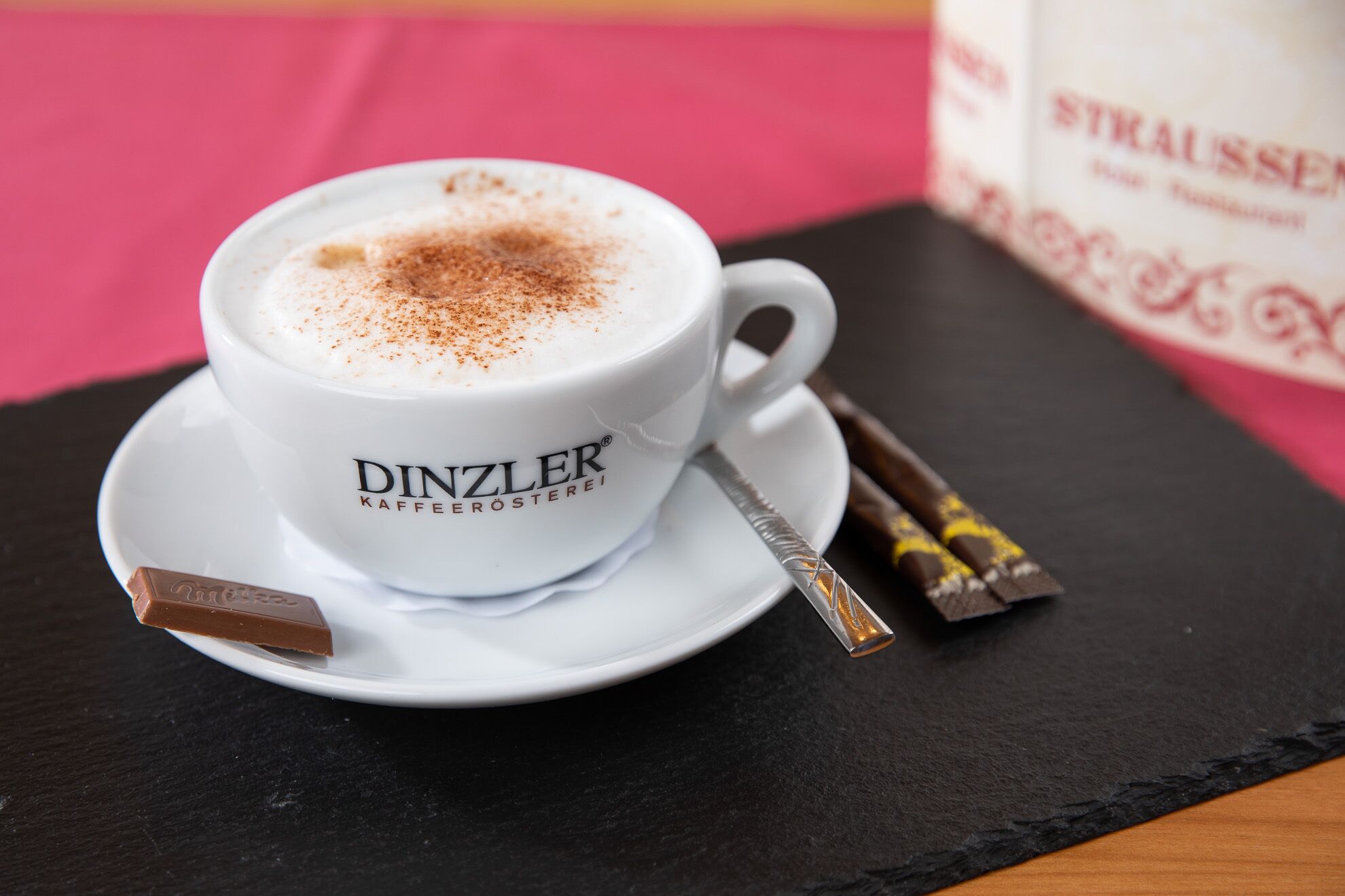A cup of coffee with a chocolate bar and a spoon on a saucer, placed on a black slate surface.