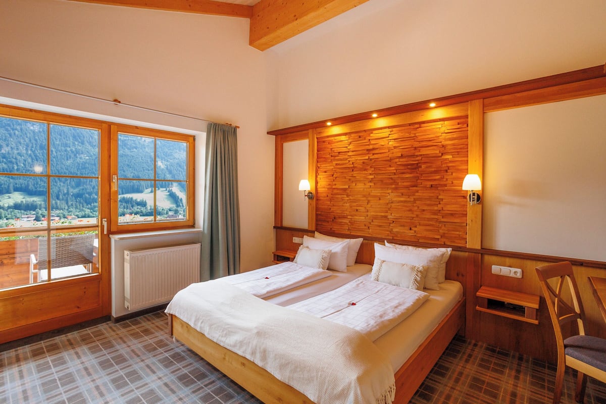 Een slaapkamer met een groot bed, een houten hoofdbord, twee bedlampen, een raam met uitzicht op een bergachtig landschap en een vloer met een ruitpatroon.