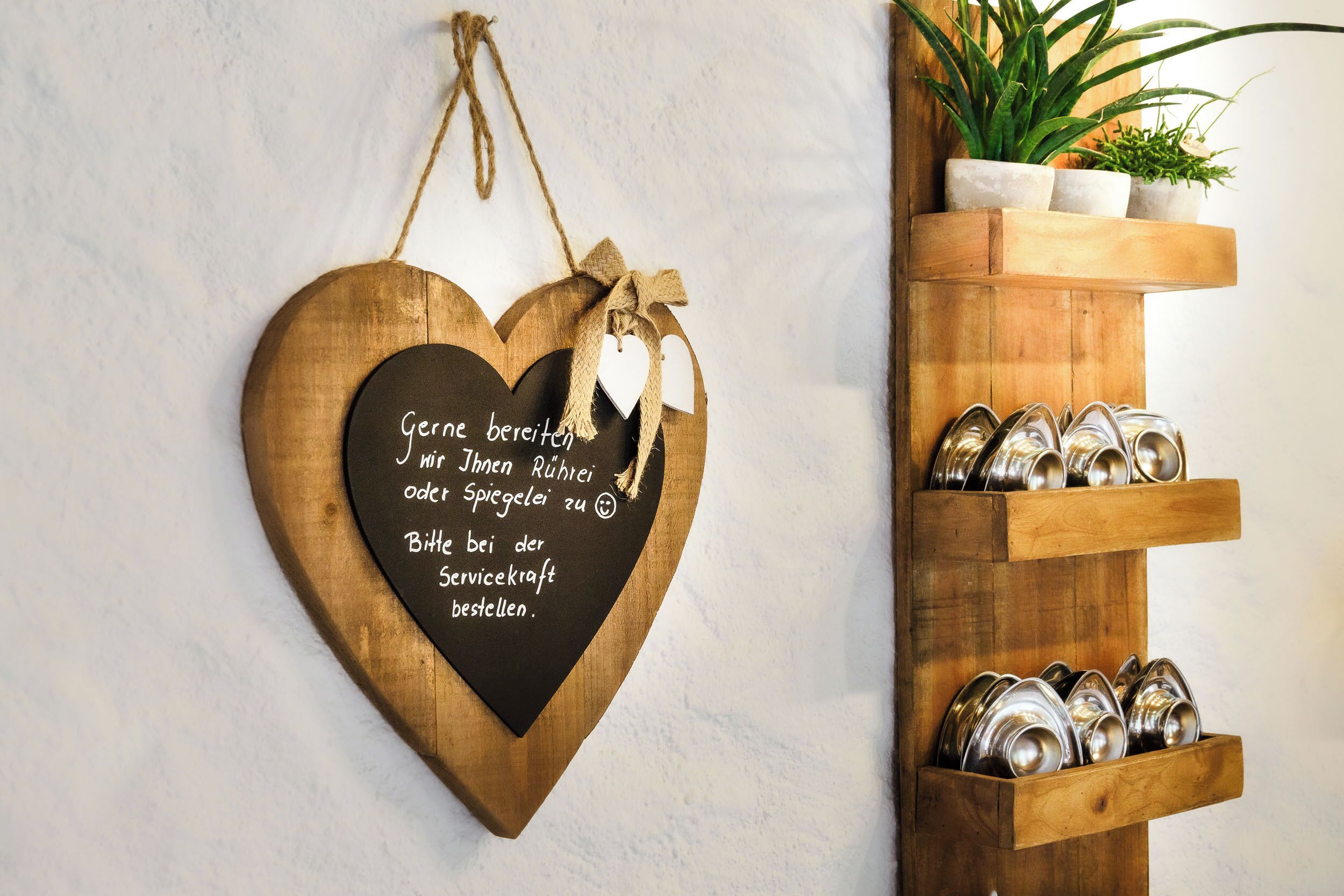 A wooden heart-shaped sign with text in German, a wooden shelf with metal dishes, and a potted plant.