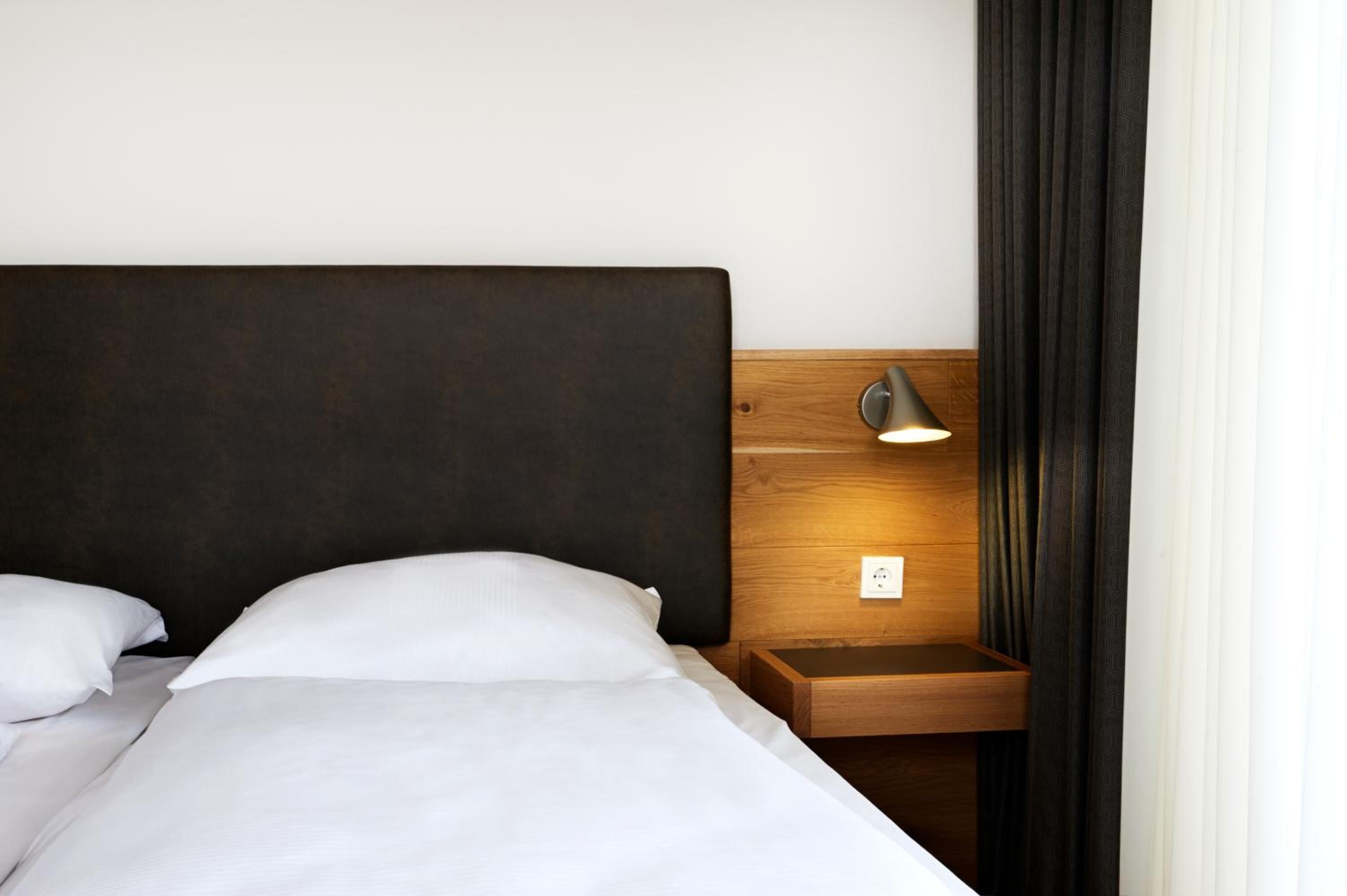 Een net opgemaakt bed met witte lakens en kussens, een donker hoofdbord, een nachtkastje met een lamp en een stopcontact.
