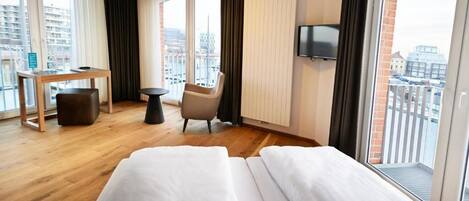 Habitación de hotel con cama, escritorio, silla, televisor y balcón con vistas a la ciudad.