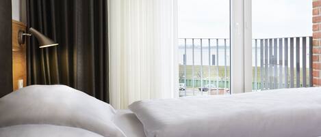 Habitación de hotel con cama, mesita de noche, lámpara y vistas a un balcón con barandilla.