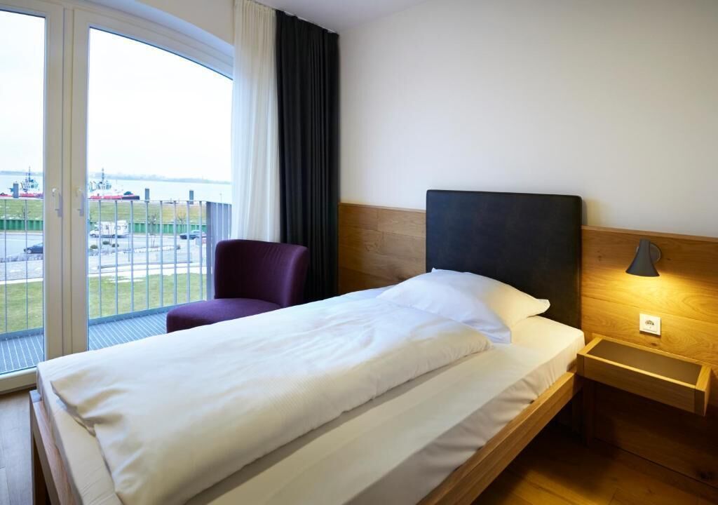 A hotel room with a bed, a chair, a nightstand, a lamp, and a view of the outside.
