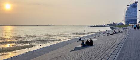 A waterfront promenade with a curved building, a pier, and a sunset.