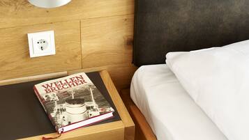 A neatly made bed with white linens, a wooden nightstand with a book titled "Wellen Brecher," and a power outlet on the wall.