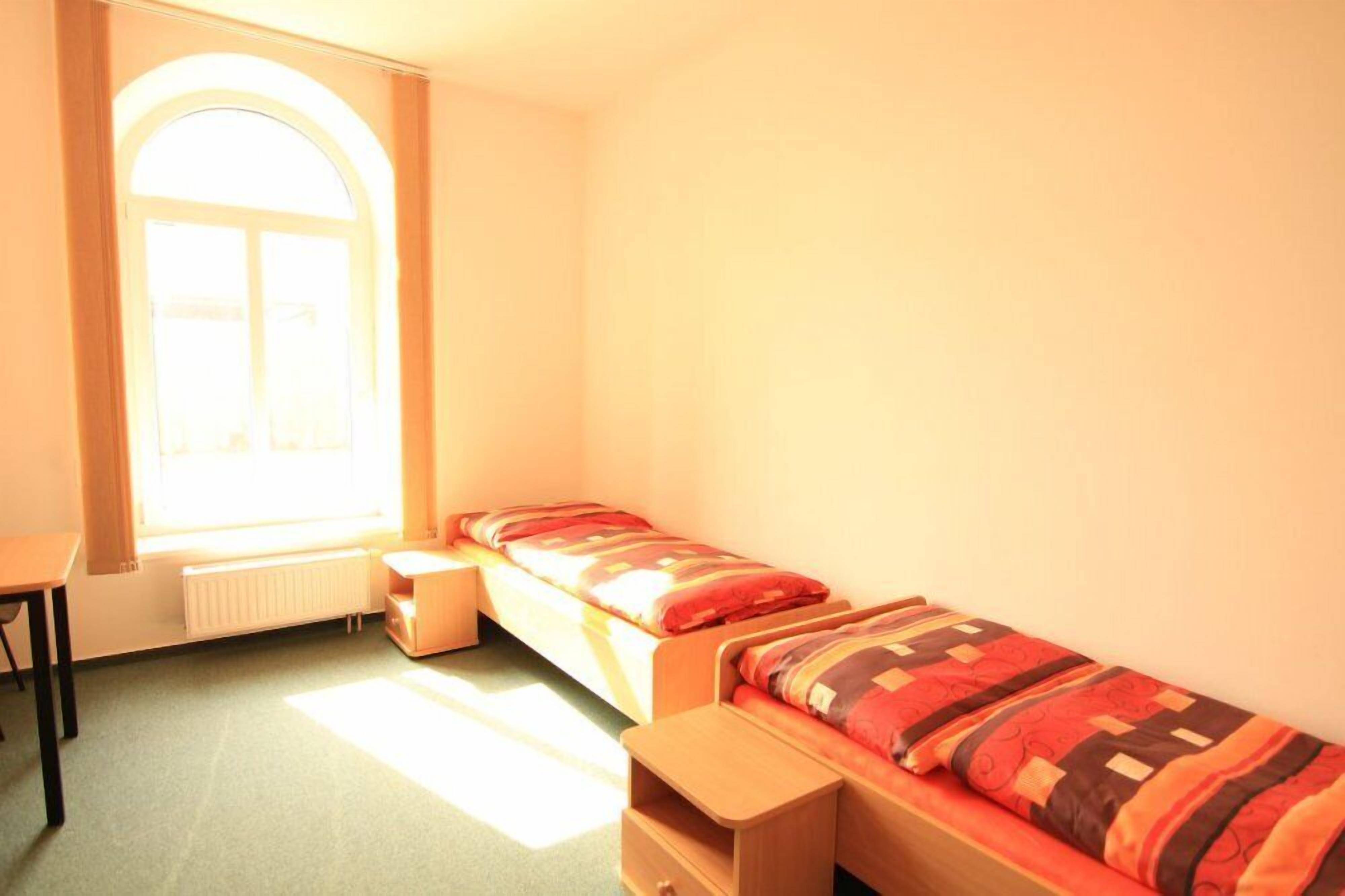 A small, bright room with two single beds, a window, and a radiator.