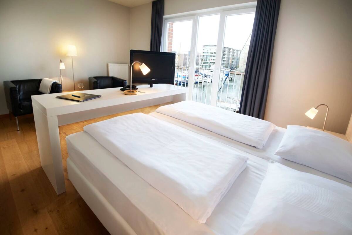 Ein Hotelzimmer mit zwei Betten, einem Schreibtisch, einem Stuhl, einem Fernseher und Blick auf die Stadt durch große Fenster.