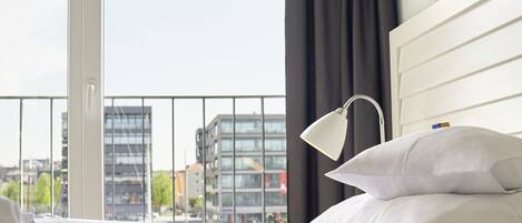 Habitación de hotel con una cama, ropa de cama blanca, una mesita de noche con lámpara y vistas a edificios a través de un ventanal grande.