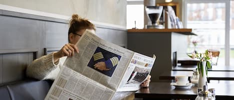 A person reading a newspaper at a table in a cafe with a view of the street outside.