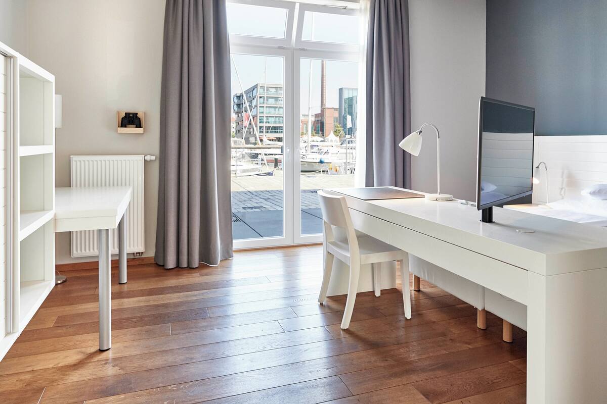 Ein modernes Hotelzimmer mit weißem Schreibtisch, einem Sessel, einem großen Fenster mit Vorhängen und Blick auf die Stadt.