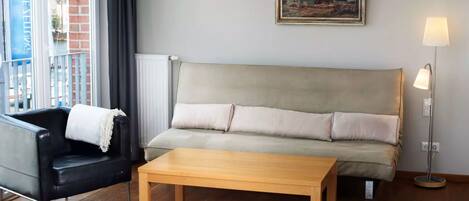 Una sala de estar con un sofá, un sillón negro, una mesa de centro de madera, una lámpara de pie y un cuadro en la pared.