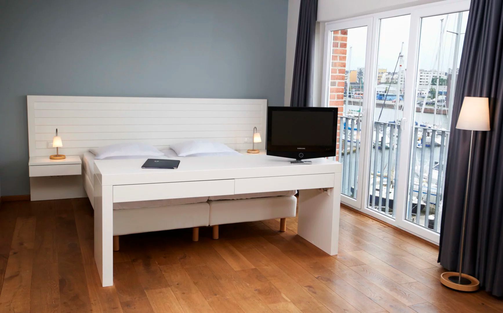 A modern hotel room with a white bed, a flat-screen TV, a desk, and a view of the marina through large windows.
