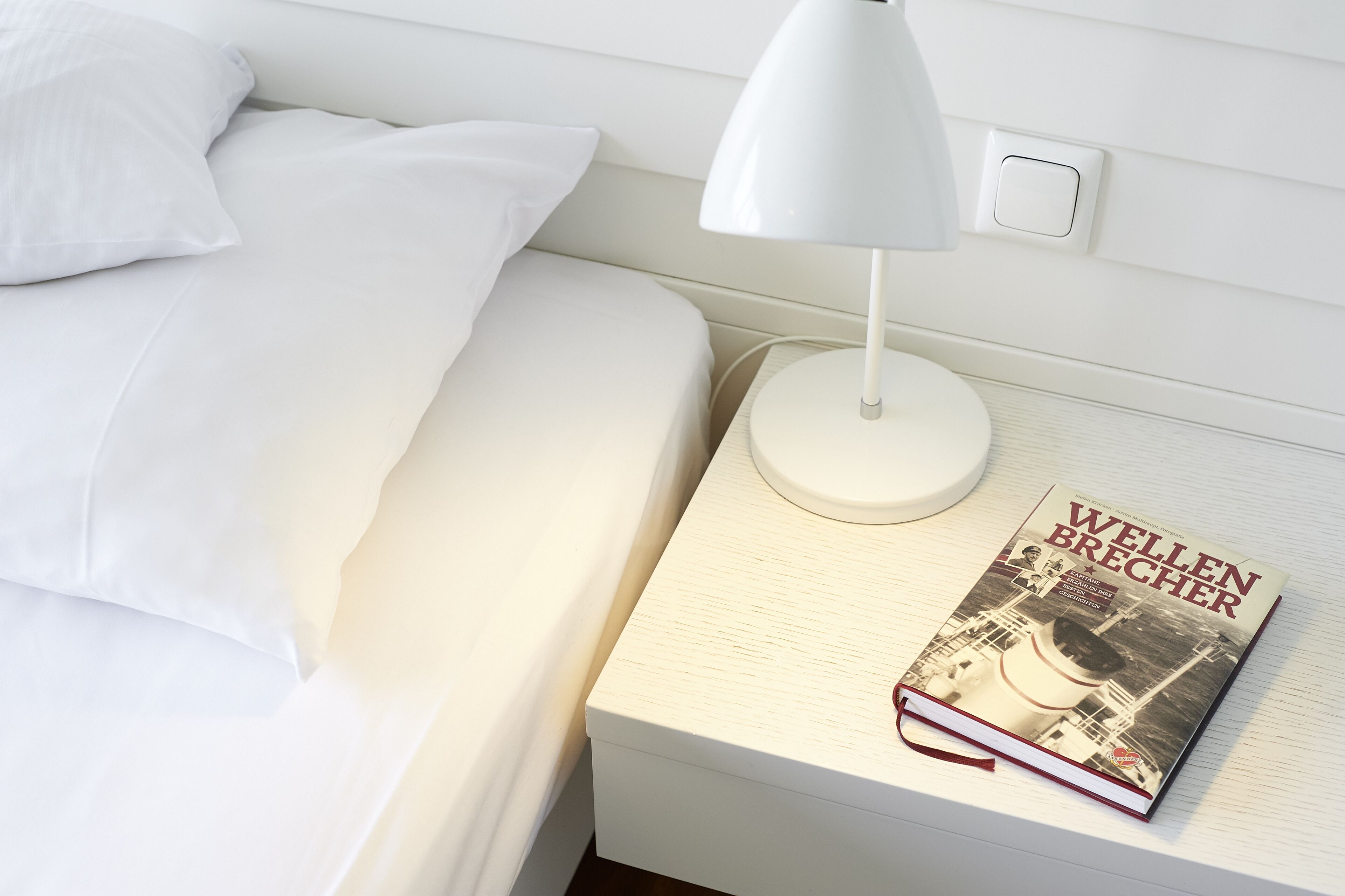 A neatly made bed with white linens, a white bedside lamp, and a book titled 'Wellen Brecher' on a bedside table.