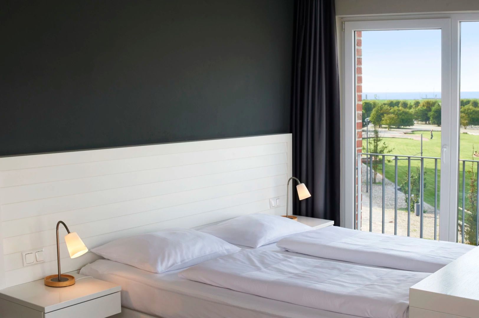 A hotel room with a bed, a nightstand, a lamp, and a view of a green landscape through a balcony.