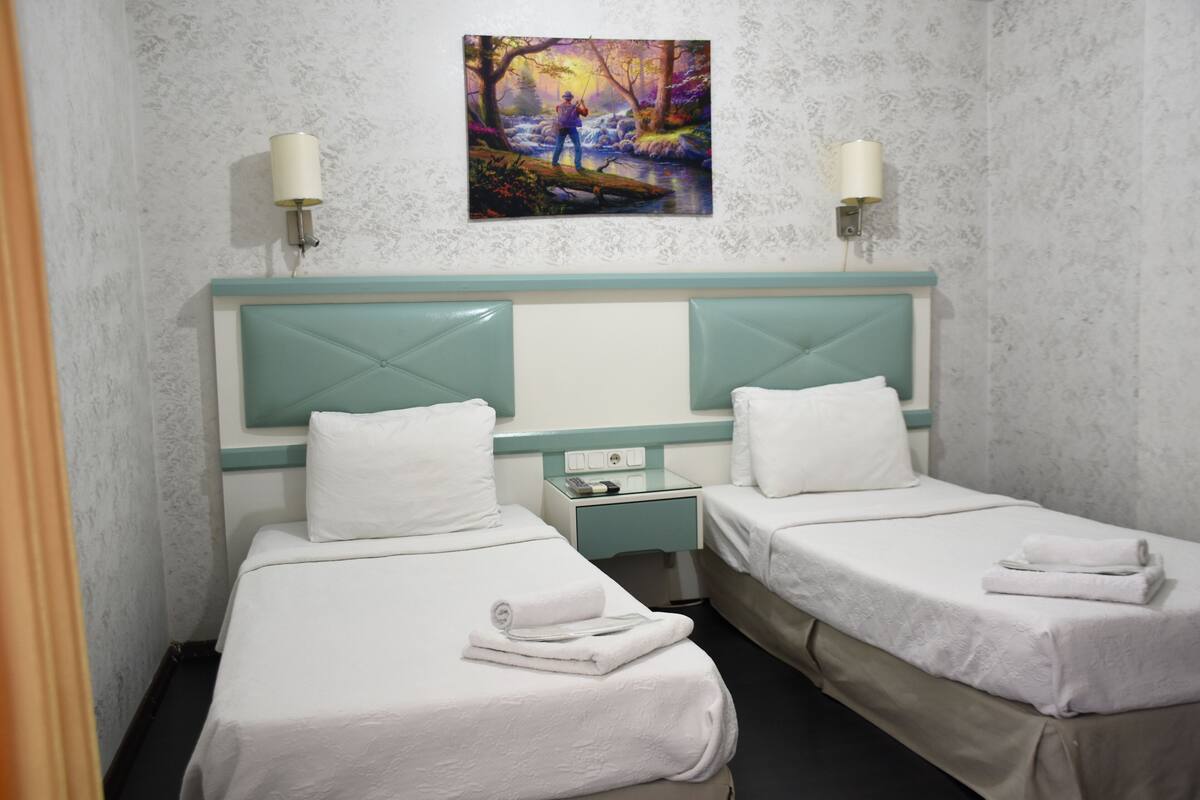 A hotel room with two single beds, a nightstand, a painting, and two wall-mounted lamps.