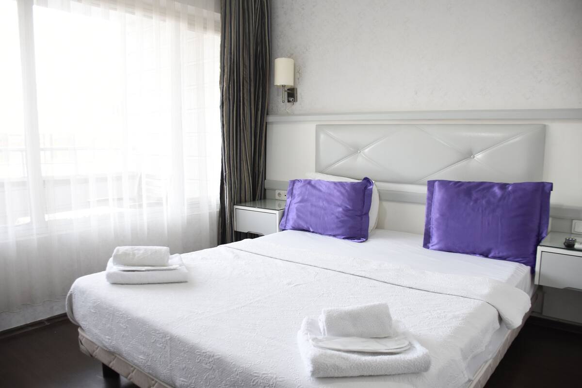 A hotel room with a large bed, white bedding, two purple pillows, a white headboard, a bedside table with a lamp, and a window with sheer curtains.