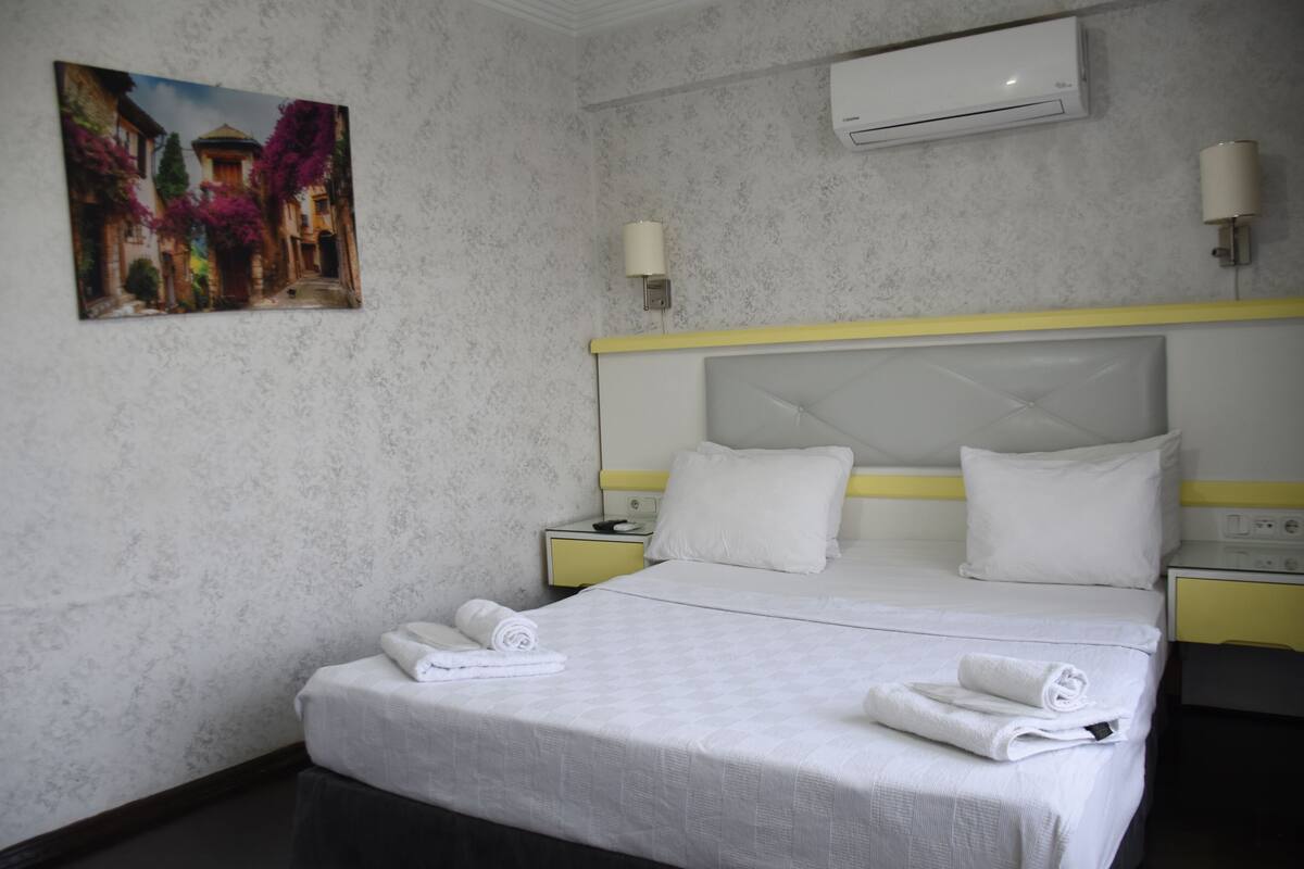 A hotel room with a bed, a painting, an air conditioner, and a nightstand.