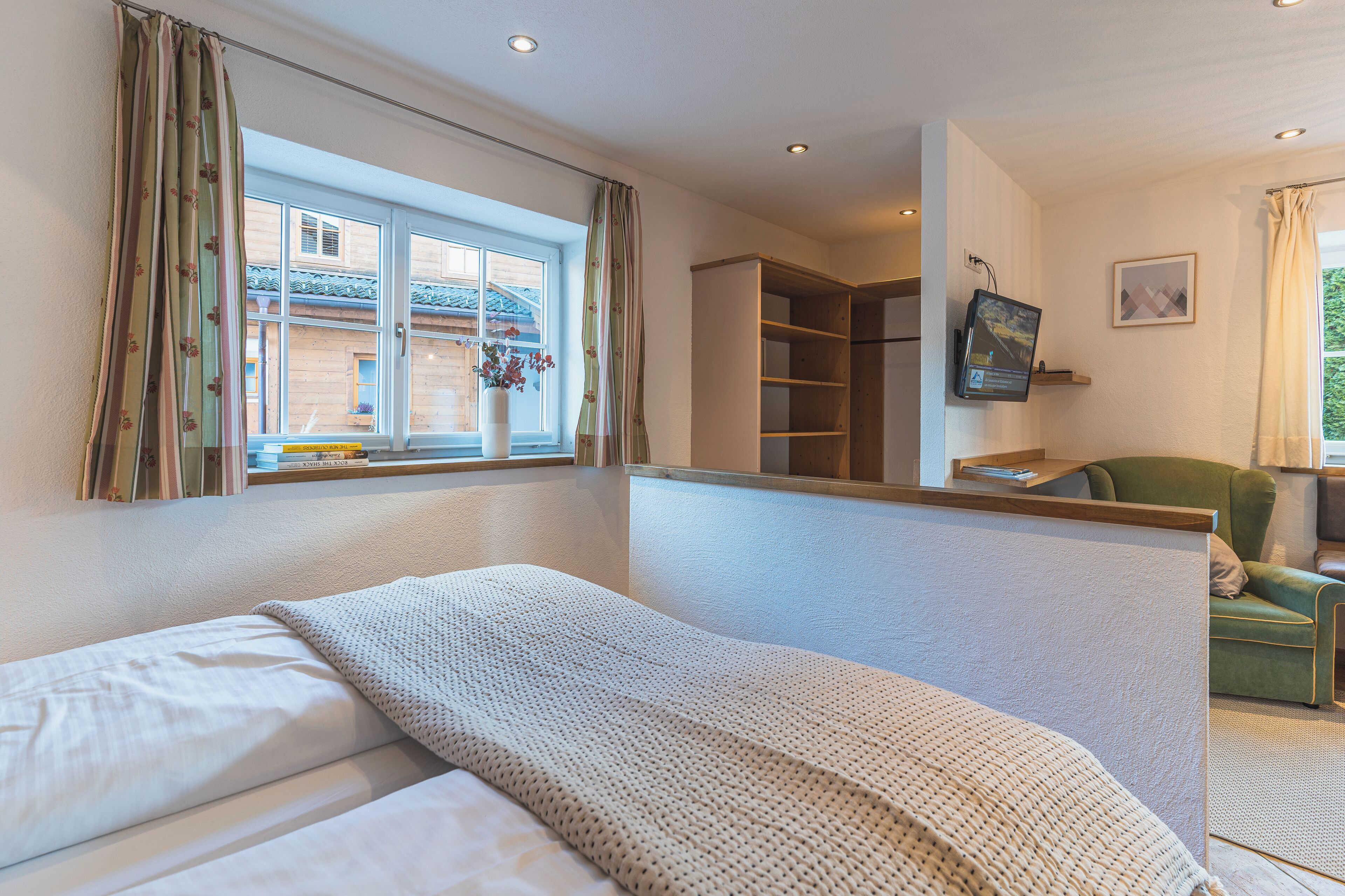 Un dormitorio con cama, ventana con cortinas, estantería y escritorio con monitor de computadora.