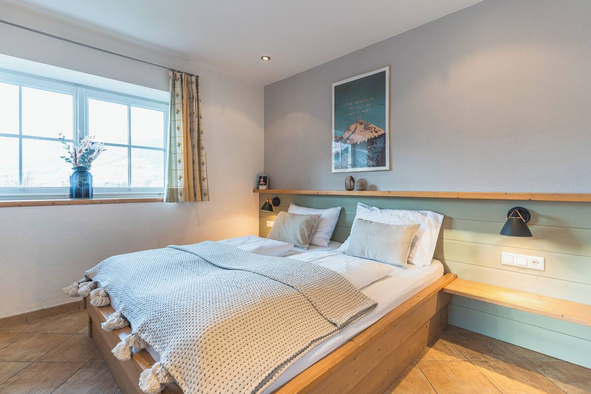 A bedroom with a wooden bed, a window with curtains, a wall-mounted lamp, and a framed picture.