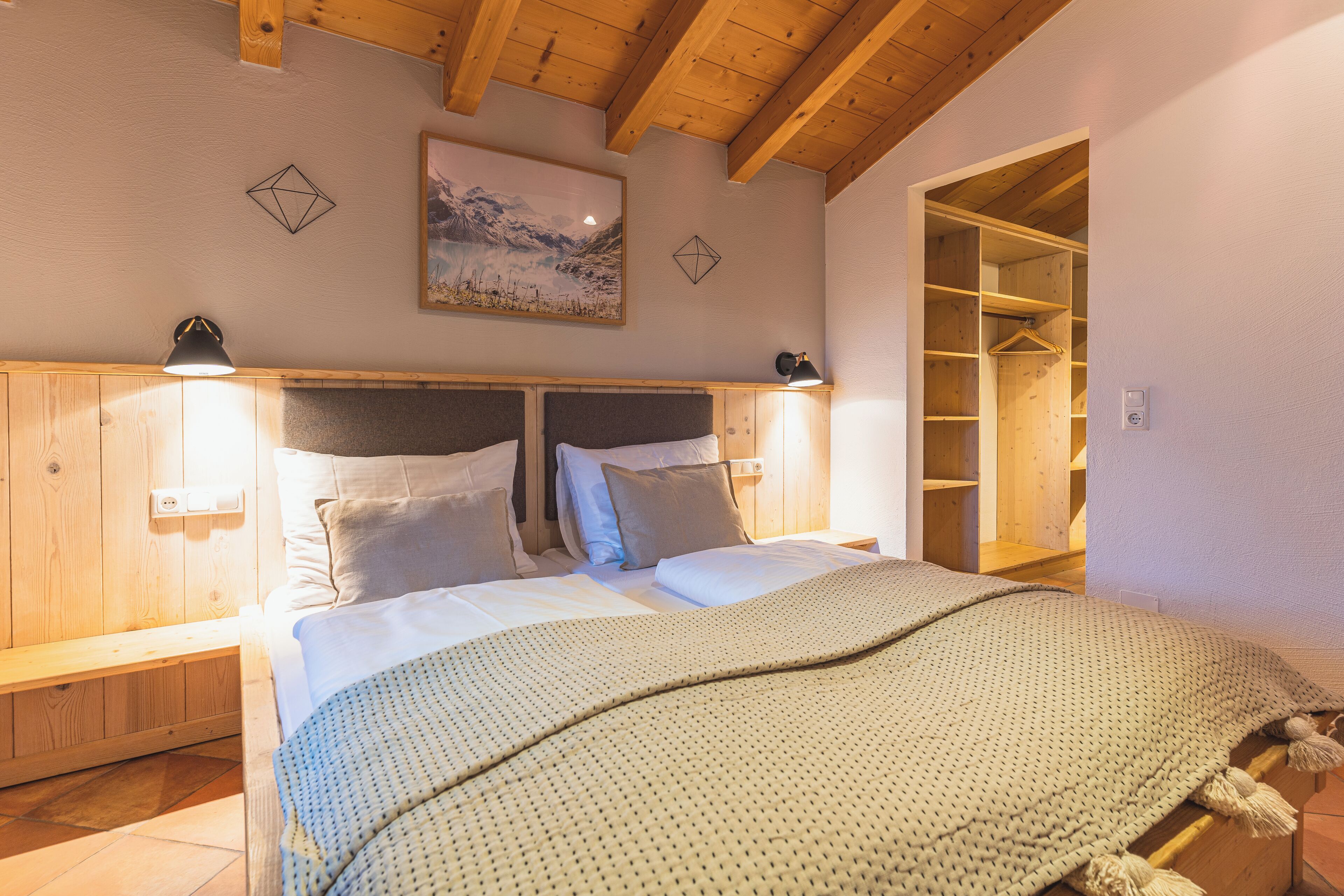 Een slaapkamer met een houten hoofdbord, twee bedlampen, een bed met witte lakens, een deken en een houten garderobe.