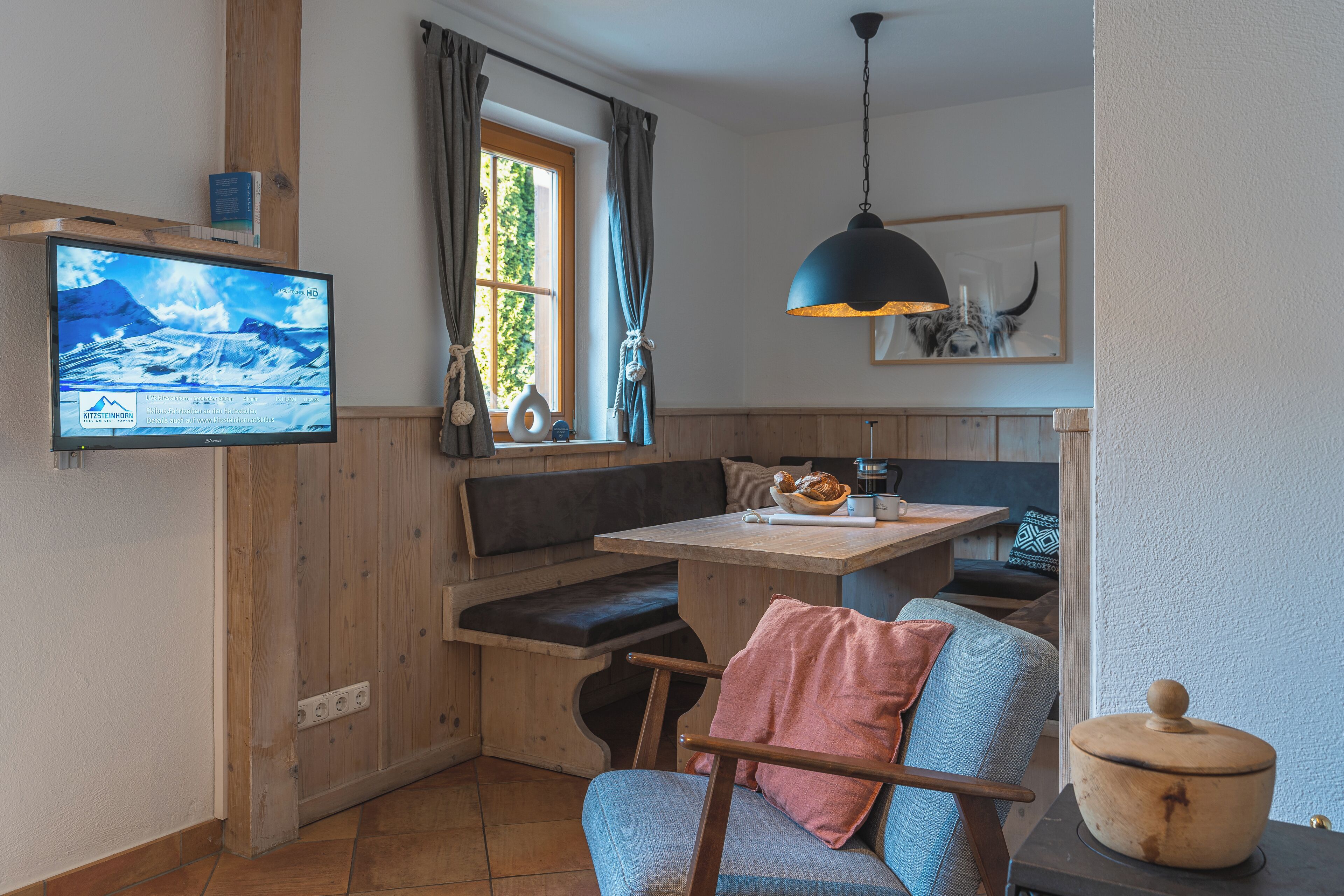 A cozy room with a wooden bench, a table, a television mounted on the wall, and a ceiling light fixture.
