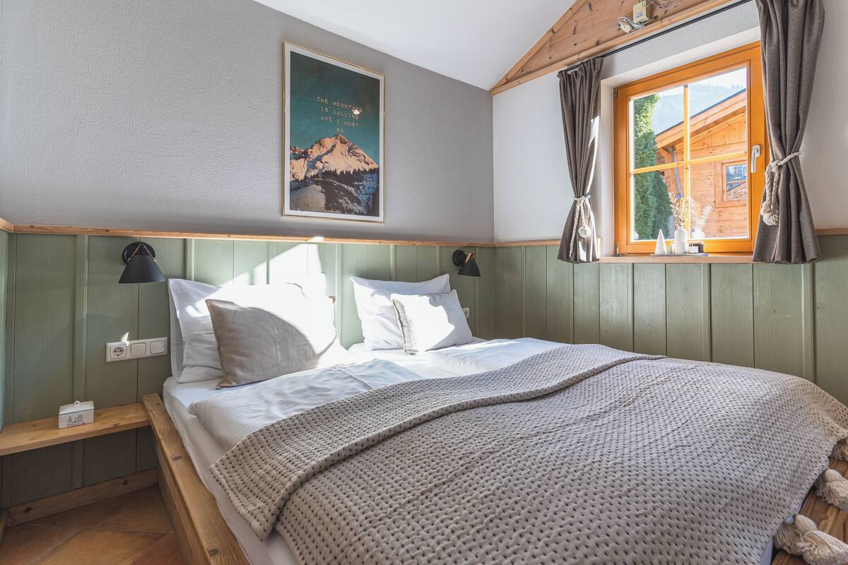 A neatly made bed with a patterned blanket, a wall-mounted lamp, and a framed picture of a mountain.