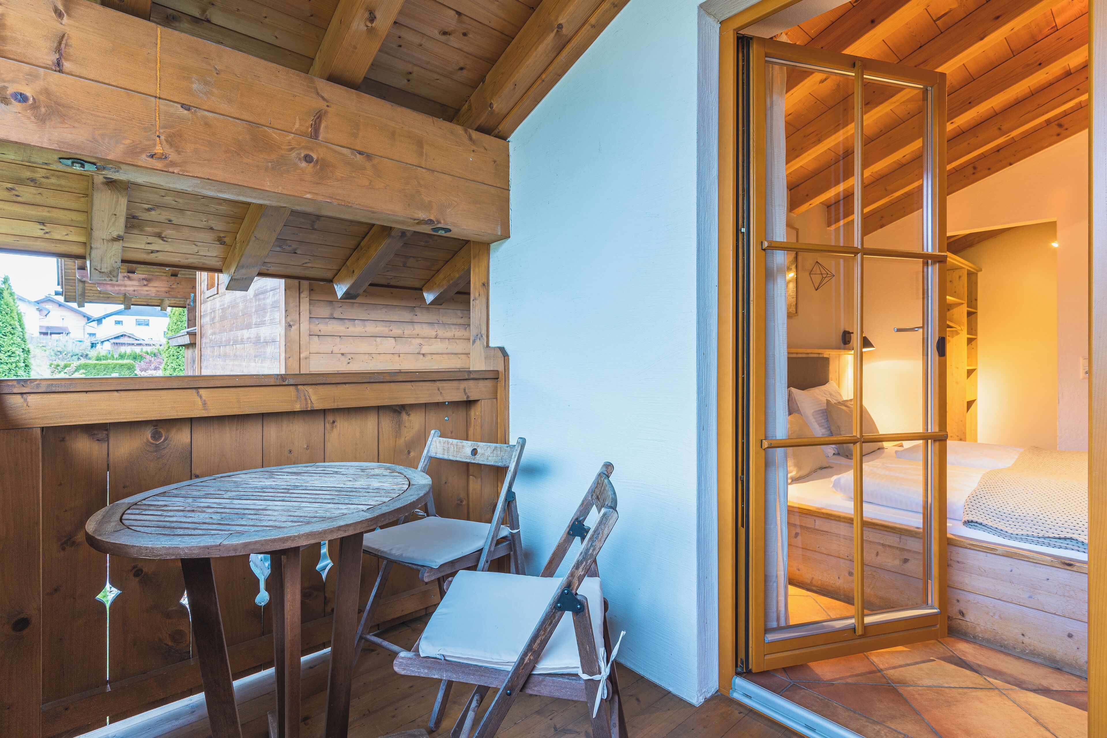 A wooden interior with a round table and chairs, a bed with a view through a glass door, and a wooden ceiling.