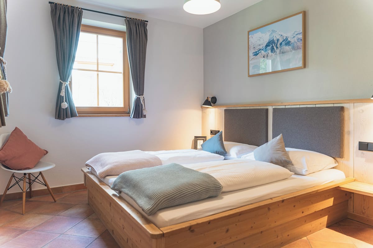 A bedroom with a wooden bed, a chair, a window with curtains, and a painting on the wall.