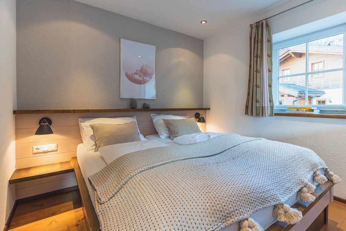 A bedroom with a bed, bedside lamps, a wall-mounted artwork, and a window with curtains.