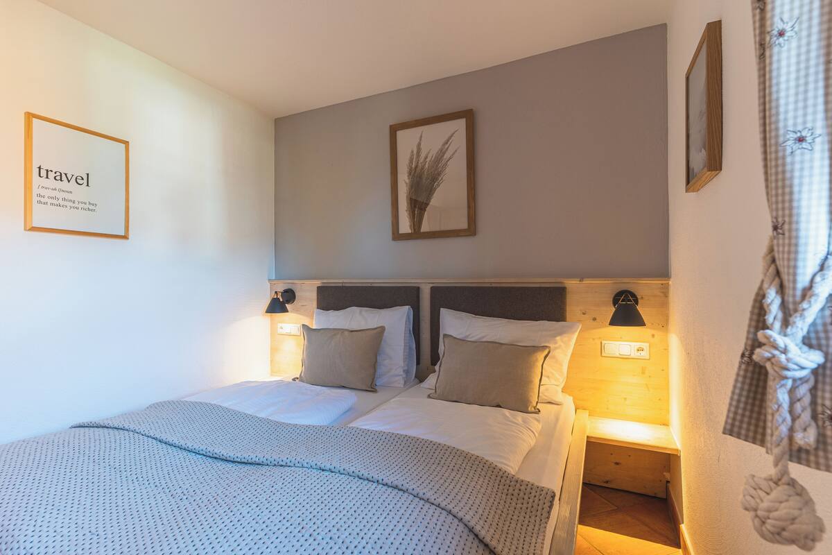 A neatly made double bed with a patterned blanket, two bedside tables with lamps, a framed quote on the wall, and a framed plant.