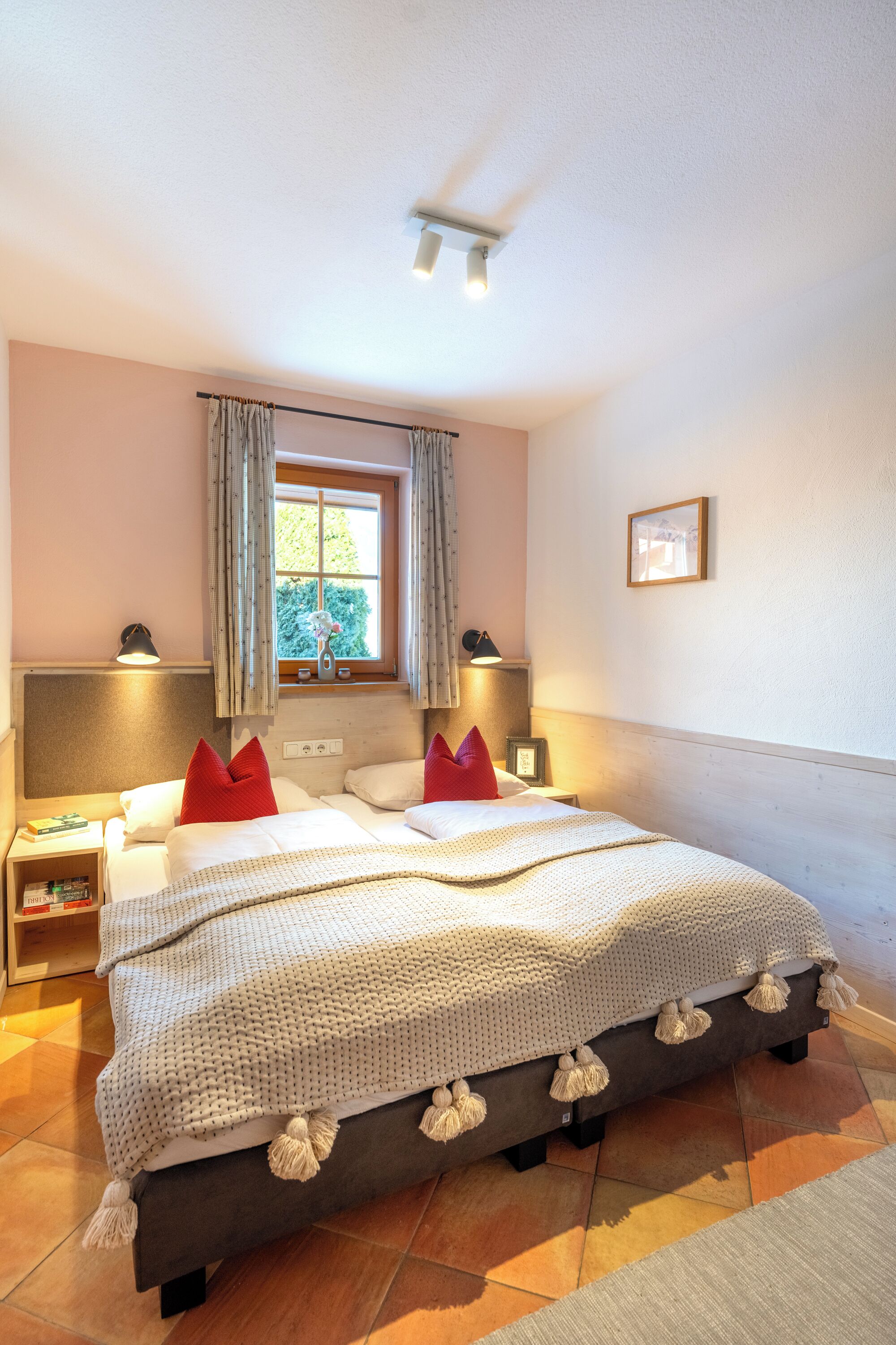 A bedroom with a bed, two bedside tables, a nightstand, a window with curtains, and a wall-mounted light fixture.